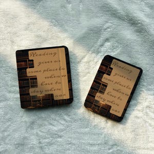 Op de afbeelding: Twee rechthoekige telefoonhoesjes met een boekthema-ontwerp. De hoesjes hebben een donkere achtergrond met een boekenplankenpatroon en een lichtgekleurde rechthoek met de tekst "Reading gives us some place to when we have to stay where oan."