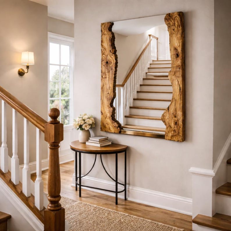 May include: A large rectangular mirror with a natural wood frame, displaying the raw edge of the wood. The mirror is mounted on a neutral wall, reflecting a staircase. A small, round table with a black metal base and a wooden top sits below the mirror.