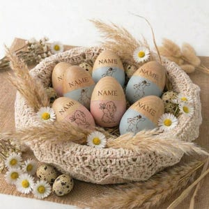 May include: A woven basket holds wooden Easter eggs with a light blue or pink painted base. Each egg has a bunny illustration and the word "NAME" engraved. The basket is decorated with daisies, dried grass, and speckled quail eggs.