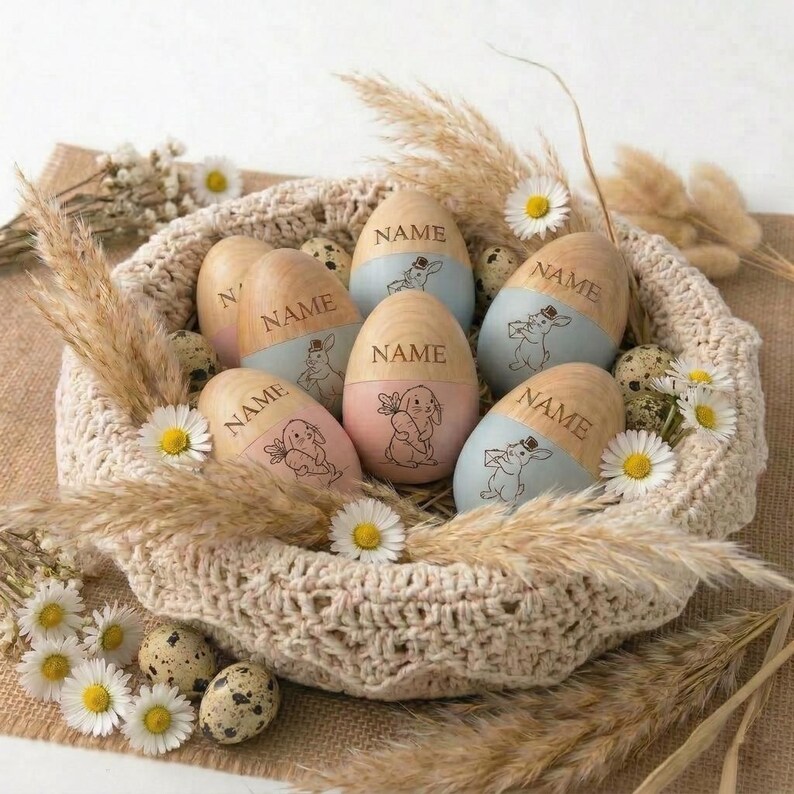 May include: A woven basket holds wooden Easter eggs with a light blue or pink painted base. Each egg has a bunny illustration and the word "NAME" engraved. The basket is decorated with daisies, dried grass, and speckled quail eggs.