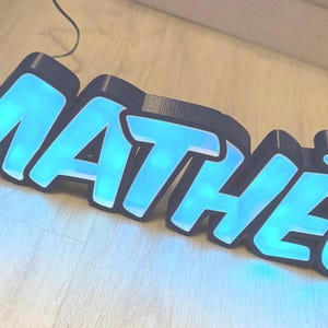 Può includere: Un cartello al neon blu con il nome "MATHEO" in lettere nere bordate. Il cartello è illuminato e si trova su una superficie di legno.