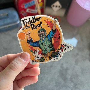 Może przedstawiać: Naklejka z kolorową ilustracją z musicalu "Skrzypek na dachu". Obraz przedstawia mężczyznę z uniesionymi ramionami na pomarańczowym tle. Naklejka ma unikalny kształt i zawiera tytuł musicalu.