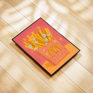 Puede incluir: Una impresión enmarcada que presenta una ilustración estilizada de papas fritas doradas en un contenedor rosa brillante con el logotipo de McDonald's. Las papas fritas están adornadas con gemas brillantes, sobre un fondo rosa y naranja.