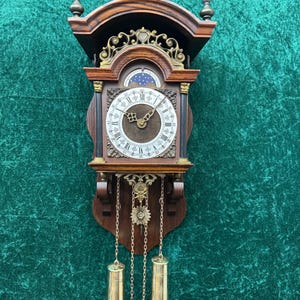 May include: A vintage wall clock with a dark wood frame, ornate gold accents, and a white clock face with Roman numerals. The clock features a pendulum and two cylindrical weights hanging below. The clock is approximately 24 inches tall.