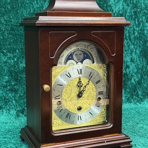 May include: A dark brown wooden mantel clock with a gold clock face and Roman numerals. The clock has a handle on top and a glass front. The clock face features a moon phase indicator.