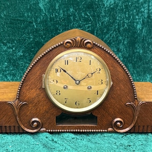 May include: A vintage wooden mantel clock with a round, gold-toned clock face. The clock has a decorative, arched top and ornate detailing. The clock's face displays black numerals and hands. The clock is approximately 12 inches wide.