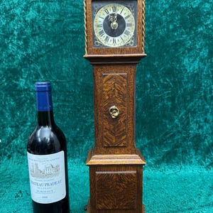 May include: A tall, antique wooden grandfather clock with a square clock face and ornate detailing. The clock has a dark wood finish and a decorative brass keyhole. A bottle of red wine with a white label is next to the clock.