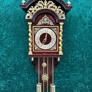 Peut inclure: Une horloge murale vintage avec un cadre en bois foncé, des accents dorés et un fond en velours rouge. Le cadran de l'horloge est blanc avec des chiffres romains noirs. Des filigranes dorés décoratifs et des poids de pendule complètent le design.