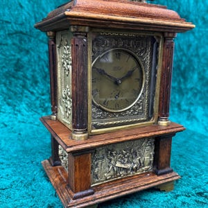 Puede incluir: Un reloj de sobremesa de madera con esfera de latón, números romanos y el texto LFS. El reloj tiene un asa de latón en la parte superior, cuatro columnas de madera oscura y un panel decorativo de latón en la base con una escena en relieve.