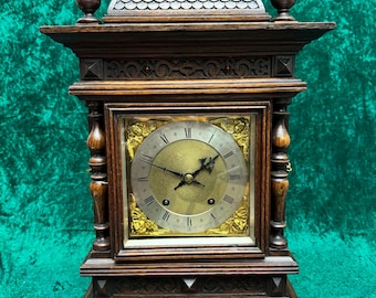 Könnte beinhalten: Eine dunkelbraune antike Uhr mit goldenem Zifferblatt. Die Uhr hat verzierte Schnitzereien und dekorative Zierknöpfe. Das Zifferblatt zeigt römische Ziffern und einen dekorativen Goldrand. Die Uhr ist aus Holz und hat eine rechteckige Form.