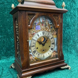 May include: A wooden antique clock with a gold-colored handle and decorative feet. The clock face features Roman numerals and a moon phase indicator. The clock's face has a painted scene with figures.