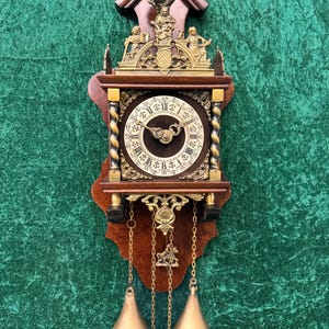 May include: A vintage wooden wall clock with a dark brown face and gold Roman numerals. The clock has decorative gold accents, a pendulum, and two brass weights. The clock is mounted on a dark brown wooden backing and has a decorative top.