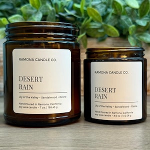 May include: Two brown glass jars of scented candles with white labels. The labels read "RAMONA CANDLE CO." and "DESERT RAIN." The larger jar contains 7 oz. of soy wax, and the smaller jar contains 3.6 oz. of soy wax. The scent includes Lily of the Valley, Sandalwood, and Ozone.