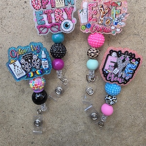 May include: Four badge reels featuring optometry-themed acrylic toppers with colorful patterns, glitter, and text including Optometry and Eye Crew. Each reel has a retractable cord and a collection of decorative beads in various colors and textures.