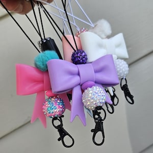 May include: A collection of colorful bow-shaped keychains. The keychains feature various colors, including pink, purple, and white, with decorative accents like pom-poms and sparkling beads. Each keychain has a black clasp for easy attachment.