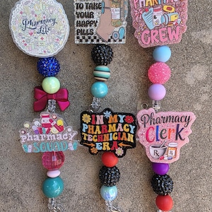 May include: Three badge reels featuring colorful acrylic toppers with pharmacy-themed graphics and text including Pharmacy Life, Don't Forget To Take Your Happy Pills, Pharmacy Crew, Pharmacy Squad, In My Pharmacy Technician Era, and Pharmacy Clerk.