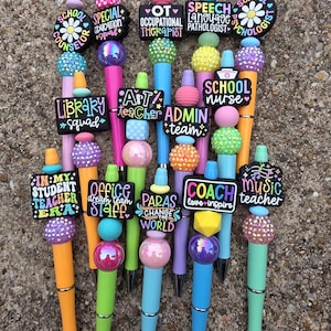 Puede incluir: Una colección de bolígrafos coloridos con adornos decorativos. Cada bolígrafo presenta un diseño diferente, con texto como "School Counselor", "OT Occupational Therapist" y "Music Teacher". Los bolígrafos tienen varios colores, incluyendo rosa, naranja y azul.
