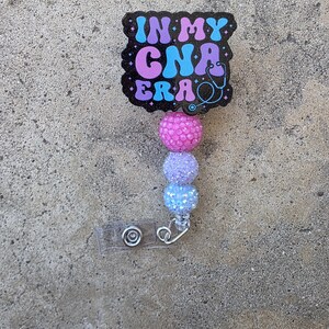 May include: A badge reel featuring a black, glittery, scalloped-edge plaque with the text IN MY CNA ERA in blue and purple, accompanied by a stethoscope icon. Below the plaque are three round, textured, glittery beads in pink, lavender, and blue.