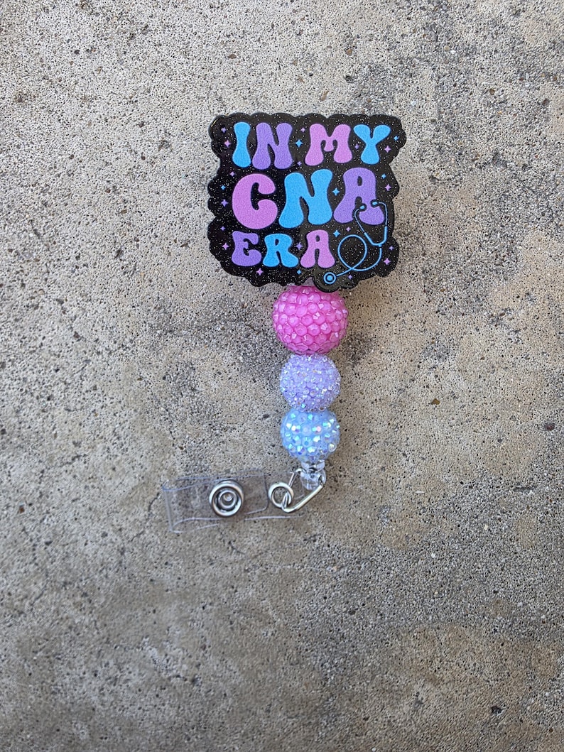 May include: A badge reel featuring a black, glittery, scalloped-edge plaque with the text IN MY CNA ERA in blue and purple, accompanied by a stethoscope icon. Below the plaque are three round, textured, glittery beads in pink, lavender, and blue.