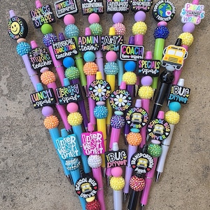 Puede incluir: Una colección de bolígrafos coloridos con adornos decorativos que muestran varios títulos y gráficos del personal escolar. Cada bolígrafo incluye cuentas texturizadas multicolores y un dije único con texto como Office Staff.
