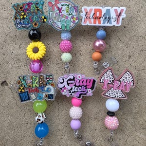 May include: A collection of six badge reels with various designs and text related to X-ray technology. The reels feature colorful beads, decorative accents, and phrases like "X-Ray Tech" and "X-Ray Technologist". The designs include images of X-ray symbols and medical equipment.