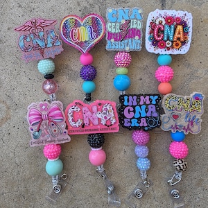 May include: Four badge reels featuring CNA designs with colorful acrylic toppers, including hearts, flowers, and medical symbols. Each reel has a beaded lanyard with various textures and colors, including pink, blue, purple, and leopard print, ending in a metal clip.