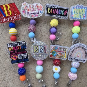 Pode incluir: Uma coleção de porta-crachás coloridos com vários designs e texto, incluindo "Therapist", "Registered Behavior Technician" e "Behavior Analyst". Cada porta-crachá apresenta contas decorativas em diferentes cores e texturas.