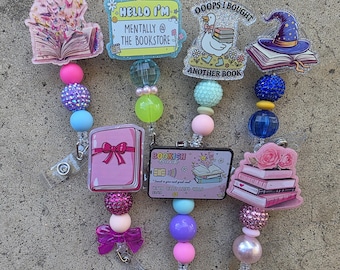 May include: Six badge reels featuring colourful acrylic charms with book-themed designs, including a duck with the text OOOPS I BOUGHT ANOTHER BOOK, a name tag reading HELLO I'M MENTALLY @ THE BOOKSTORE, and a credit card labelled BOOKISH BANK.