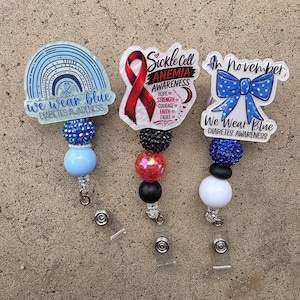 May include: Three badge reels featuring awareness themes. The left shows a blue rainbow with text "In November we wear blue Diabetes Awareness". The center shows a red ribbon with "Sickle Cell Anemia Awareness Hope Strength Courage Faith Fight". The right shows a blue bow with "In November We Wear Blue Diabetes Awareness". Each has colorful beads.