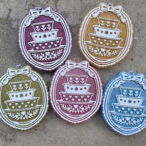 May include: Five oval-shaped hanging ornaments in purple, gold, green, pink, and blue. Each features a white embossed design of a four-tiered stack of bowls decorated with hearts and stars, framed by a white border with a bow at the top.