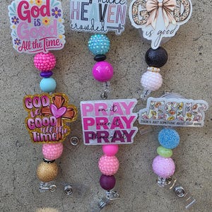 May include: Three badge reels featuring acrylic toppers with religious text and designs, including God is Good, Make Heaven Crowded, and Jesus. Each reel is decorated with colorful, textured, and glittery beads in pink, blue, gold, and black tones.