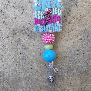 May include: A badge reel featuring a rectangular glittery acrylic topper with the text CNA CERTIFIED NURSING ASSISTANT and a pink leopard print lightning bolt. Below the topper are three beads in pink, light green, and blue, with a clear strap.