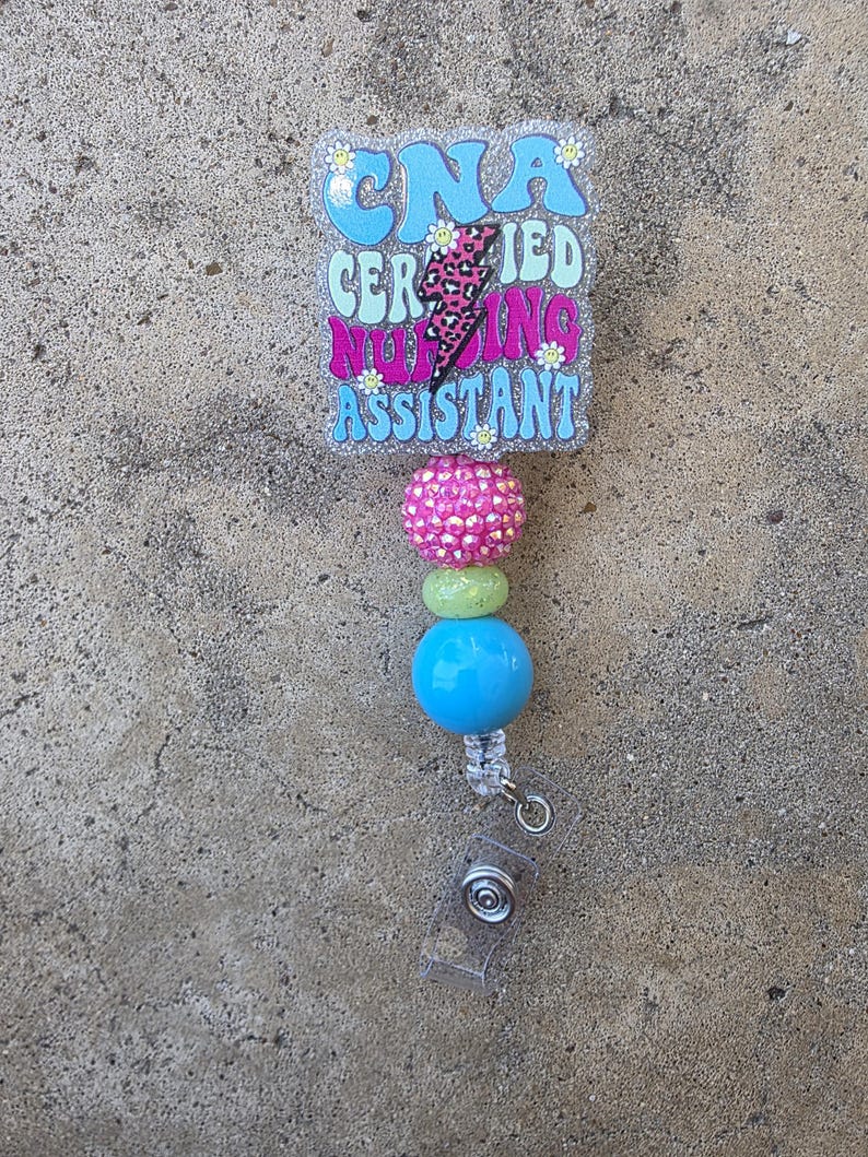 May include: A badge reel featuring a rectangular glittery acrylic topper with the text CNA CERTIFIED NURSING ASSISTANT and a pink leopard print lightning bolt. Below the topper are three beads in pink, light green, and blue, with a clear strap.