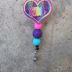 May include: A heart-shaped badge reel featuring a rainbow leopard print pattern and the letters CNA in a white script font. The design is accented with a pink glitter border and a vertical strand of three beads in pink, purple, and light blue.