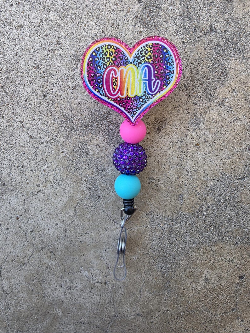 May include: A heart-shaped badge reel featuring a rainbow leopard print pattern and the letters CNA in a white script font. The design is accented with a pink glitter border and a vertical strand of three beads in pink, purple, and light blue.
