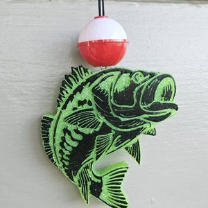 May include: A hanging ornament shaped like a fish, featuring a black and neon green glittery finish. The fish is suspended from a black cord attached to a round, red and white plastic fishing bobber that sits directly above the fish figure.