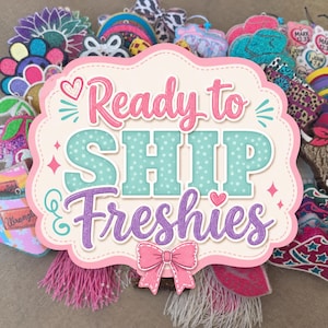 May include: A collection of colorful, glittery, and patterned hanging air fresheners, known as freshies, arranged behind a central sign that reads Ready to SHIP Freshies in pink, teal, and purple text with a pink bow graphic.