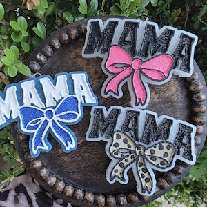 May include: Three decorative ornaments spelling "MAMA" with bow accents. One has a pink bow, one a blue bow, and the third a leopard print bow. The ornaments are white with black or blue lettering and are displayed on a wooden tray.