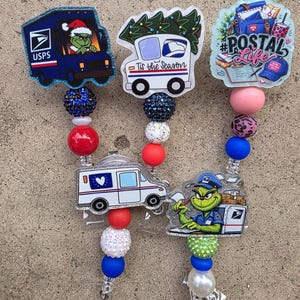 May include: Five badge reels featuring USPS and holiday themes. Designs include a Grinch-driven mail truck, a truck with a tree and 'Tis the Season' text, a '#POSTAL Life' graphic, a white mail truck, and a Grinch in a postal uniform.