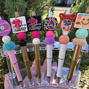 May include: A collection of decorative pens with various toppers. The toppers include a smiley face, a lightbulb, and inspirational quotes like "She is Strong" and "I am Worthy." The pens have colorful accents and are displayed in a clear holder.