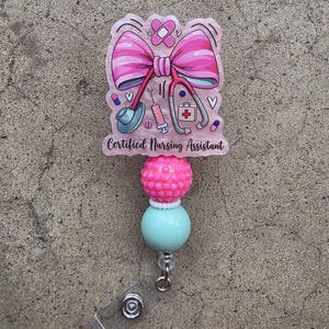 May include: A badge reel featuring a pink acrylic topper with a large pink bow, a stethoscope, a syringe, a first aid kit, and a bandage. The text "Certified Nursing Assistant" is printed in black. Below the topper are one textured pink bead and one smooth mint green bead.