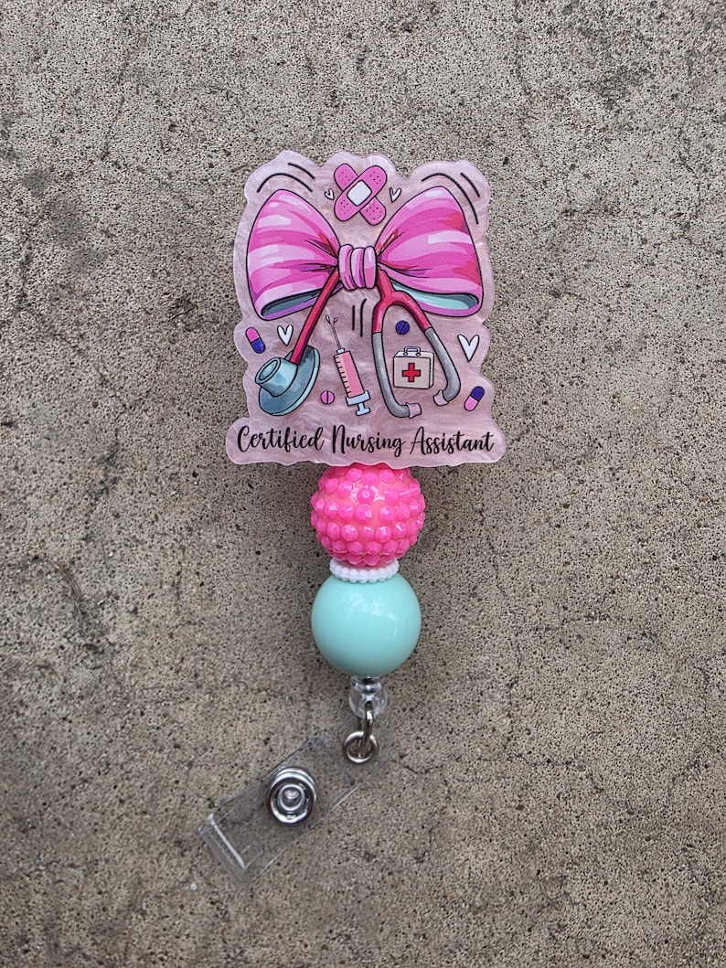 May include: A badge reel featuring a pink acrylic topper with a large pink bow, a stethoscope, a syringe, a first aid kit, and a bandage. The text "Certified Nursing Assistant" is printed in black. Below the topper are one textured pink bead and one smooth mint green bead.
