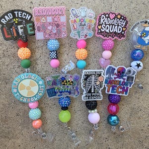 Peut inclure: Cinq enrouleurs de badge avec des décorations en acrylique sur le thème de la radiologie et des cordons perlés colorés. Les motifs incluent des textes comme RAD TECH LIFE, RADIOLOGY SQUAD et RADIOLOGY TECH, avec des squelettes et symboles.