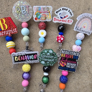 Puede incluir: Una colección de carretes de insignia coloridos con varios diseños y texto, incluyendo "Therapist", "RBT" y "Behaviors". Cada carrete presenta una combinación única de cuentas en diferentes colores y formas.