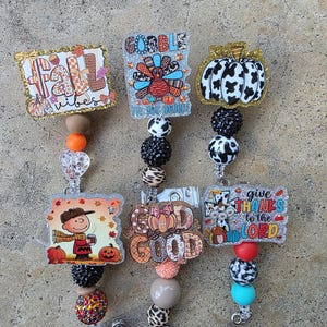 May include: Three badge reels featuring decorative acrylic toppers with fall and Thanksgiving themes, including text like fall vibes, gobble, and give thanks to the lord. Each reel has a beaded lanyard with various colors, patterns, and textures.