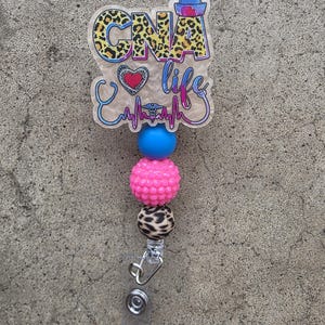 May include: A badge reel featuring a translucent acrylic topper with the text CNA life, a leopard print pattern, a heart, and a stethoscope graphic. Below the topper are three beads in blue, textured pink, and leopard print, ending in a metal clip.
