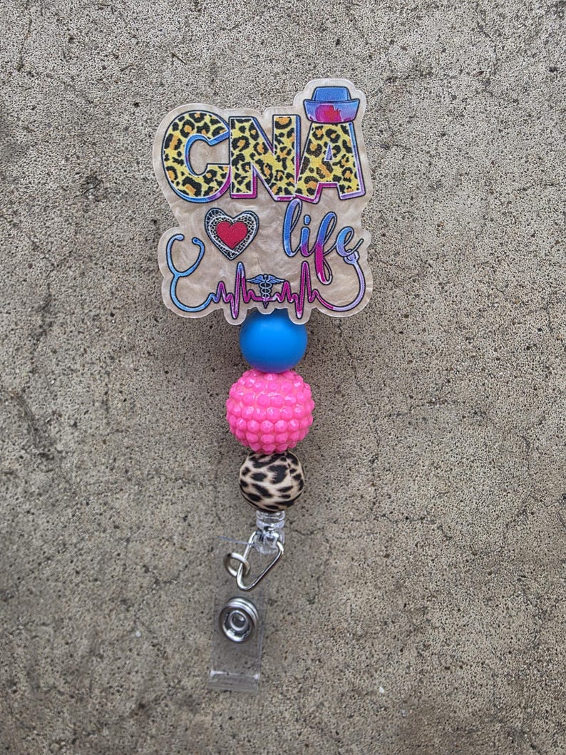 May include: A badge reel featuring a translucent acrylic topper with the text CNA life, a leopard print pattern, a heart, and a stethoscope graphic. Below the topper are three beads in blue, textured pink, and leopard print, ending in a metal clip.
