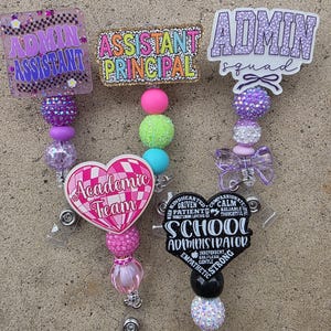 May include: Four badge reels featuring decorative acrylic toppers and colorful beaded lanyards. Toppers include text reading Admin Assistant, Assistant Principal, Admin squad, Academic Team, and School Administrator with various descriptive words.