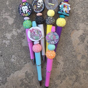 Puede incluir: Una colección de cinco bolígrafos decorativos con cuentas de colores y adornos temáticos. Los adornos incluyen diseños como un diente con el texto Dental Squad, una calavera con Slingin-Pills, gráficos de X-Ray tech y Rainbows & Butterflies.