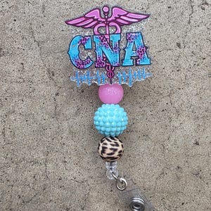May include: A badge reel featuring a glittery acrylic topper with a pink caduceus symbol, the text CNA in blue and purple leopard print, and a blue heartbeat line. Below the topper are three beads: one pink, one textured light blue, and one leopard print.